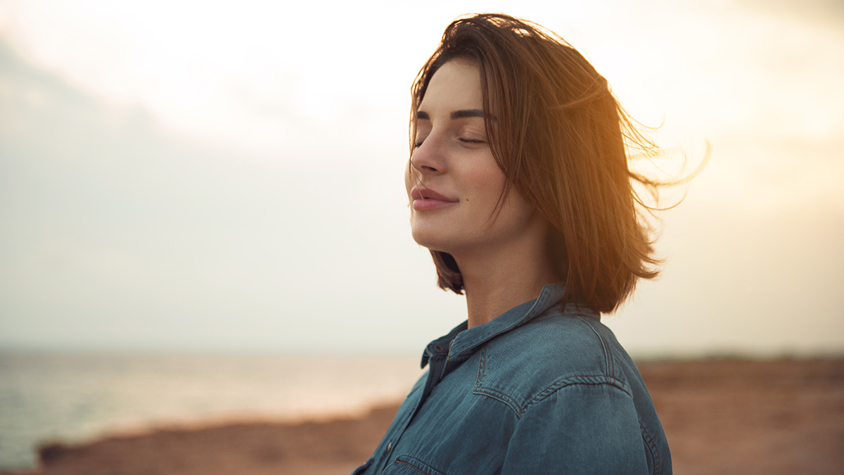 Beautiful and content dark-haired woman on the beach in a light jeans shirt with the bright sun behind her back and her eyes closed as she smiles